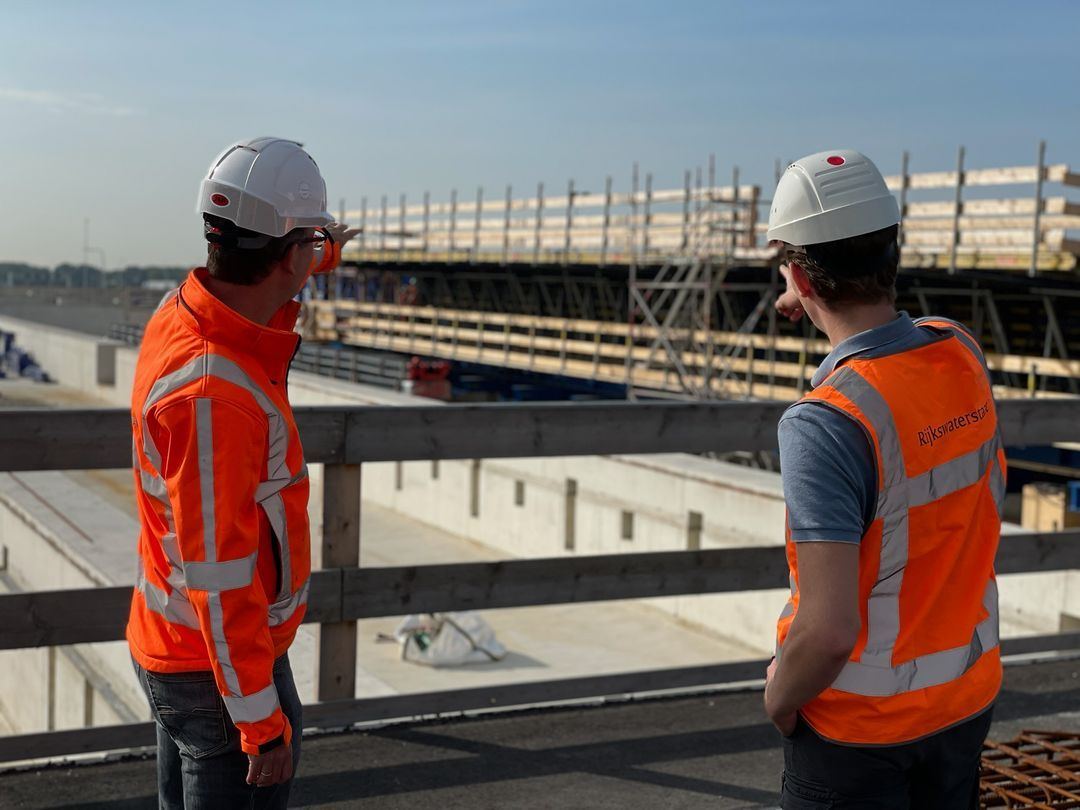 Twee collega's in oranje hesjes en witte helmen kijken uit over een bouwplaats aan een brug